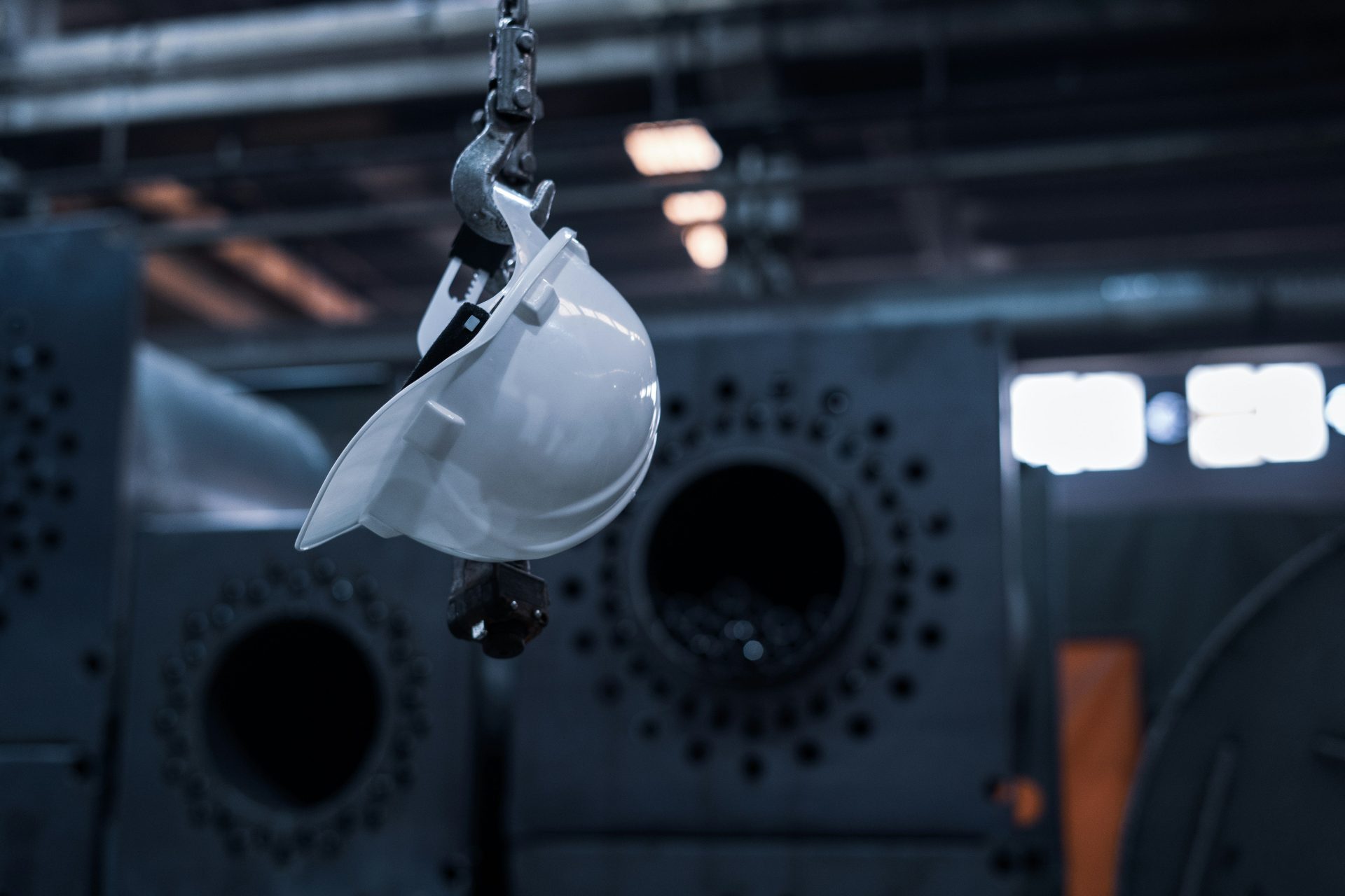 A white hard hat hanging from a metal hook in a dark industrial factory
